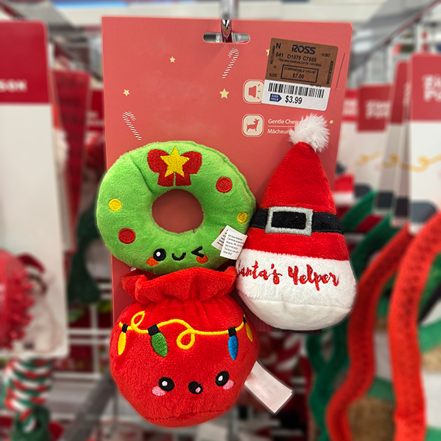 Set of three holiday-themed plush dog toys hanging on a store display: a green wreath with a smiling face, a red gift bag decorated with Christmas lights, and a Santa hat labeled “Santa’s Helper.”