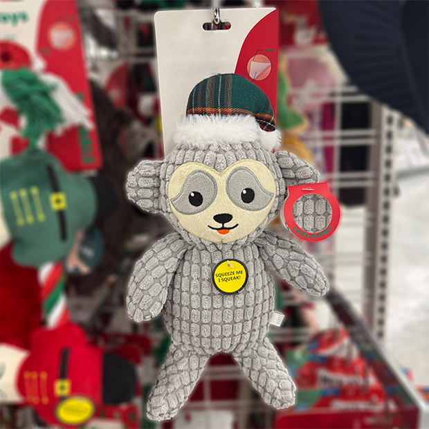 Gray textured plush dog toy with floppy limbs wearing a plaid winter hat, displayed on a store shelf with a tag that reads “Squeeze Me I Squeak.”