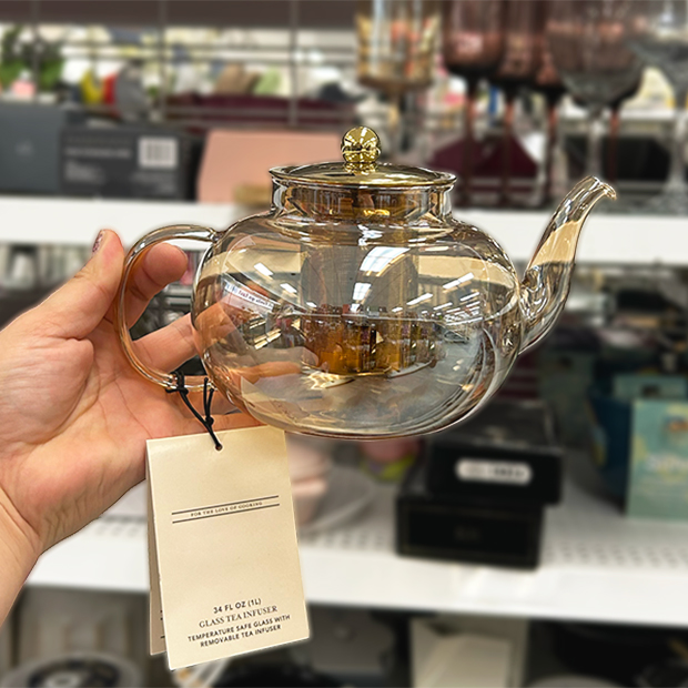 Clear glass teapot with a gold-colored lid and infuser, held up in store at Ross.
