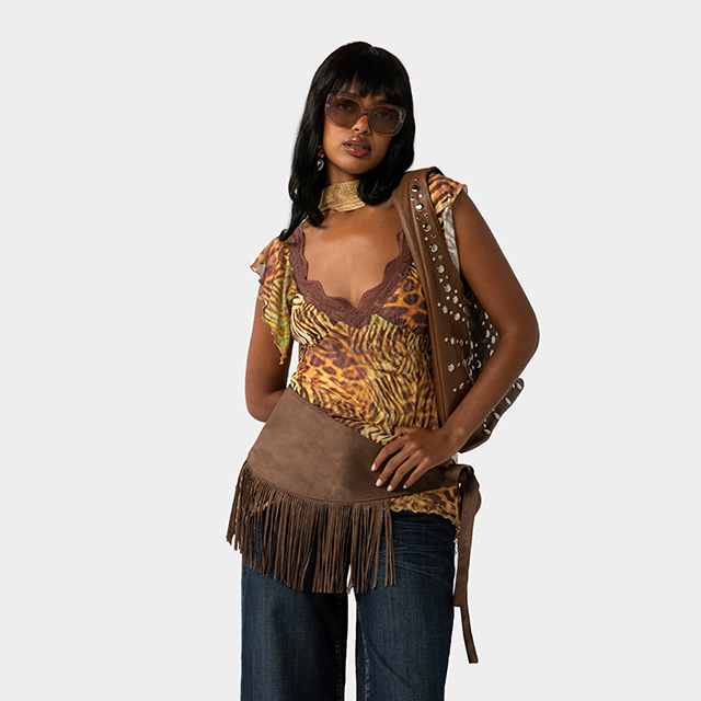 Model wears animal printed top with lace trim and jeans with a fringed scarf around the waist and studded handbag and large sunglasses