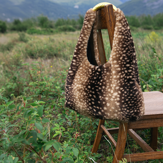 Doe-print handbag draped over a wooden chair in a grassy field with mountains in the background.