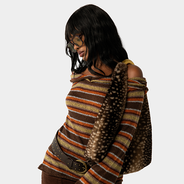 Model wearing an off-the-shoulder striped knit sweater, brown pants, and a studded belt, accessorized with oversized sunglasses and an animal-print shoulder bag for a retro-inspired fall look.