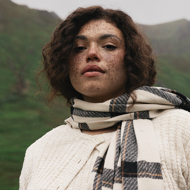 Close-up of a person wearing a cream knit sweater and plaid scarf outdoors against a green mountain backdrop.