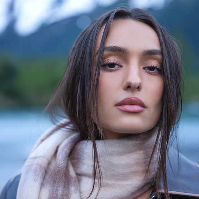 Close-up of a person wearing a soft plaid scarf and leather jacket outdoors with a blurred mountain and lake backdrop.